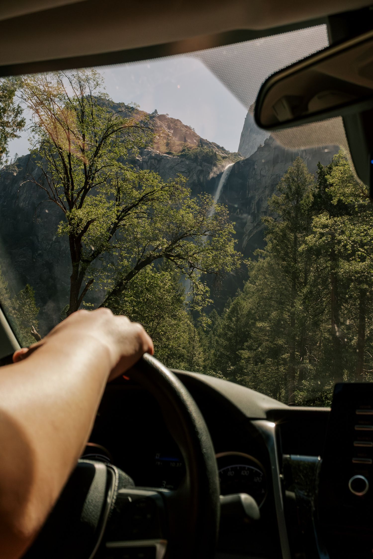 View from inside a car driving through Yosemite National Park with a waterfall and mountain peak visible through windshield.