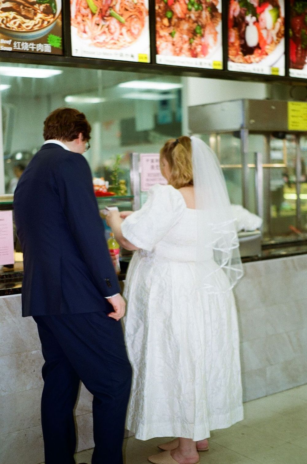 A couple in wedding attire ordering food at a casual restaurant counter with menu boards displayed above.