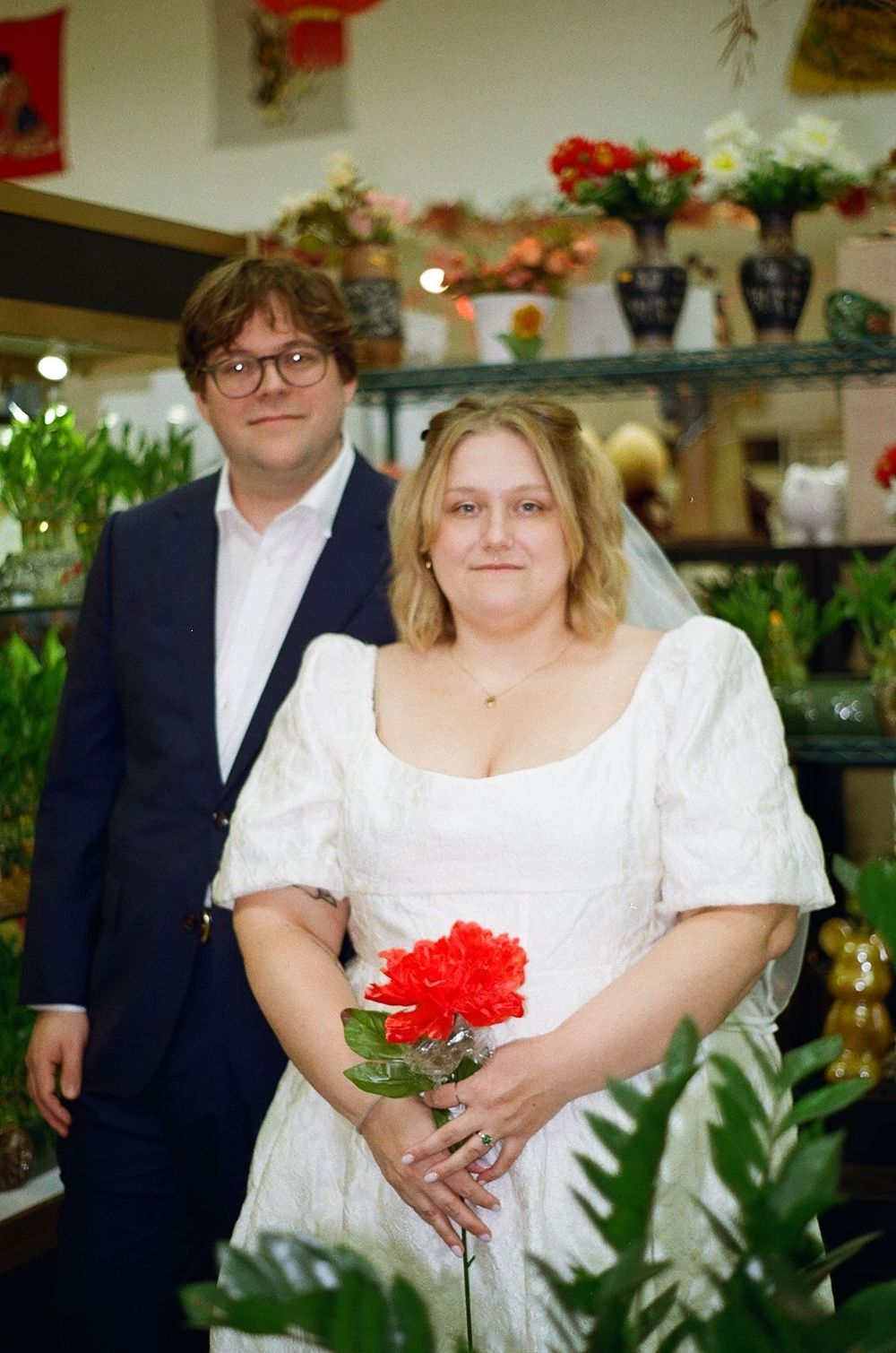 A bride in a white dress holds a red carnation while standing in a flower shop with floral displays on shelves behind her.