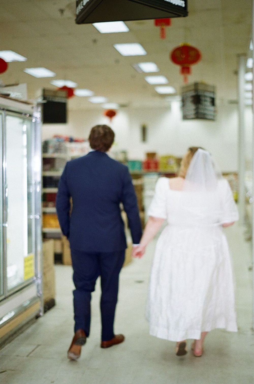 A couple in wedding attire walks down a retail aisle decorated with red lanterns.