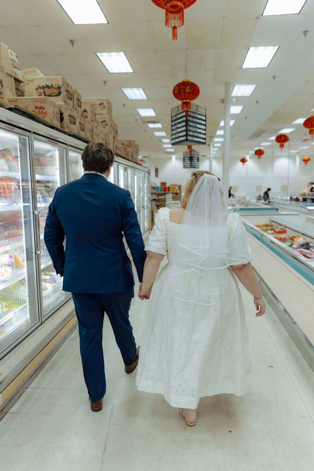 A couple in wedding attire walk through a Chinese grocery store decorated with red lanterns.