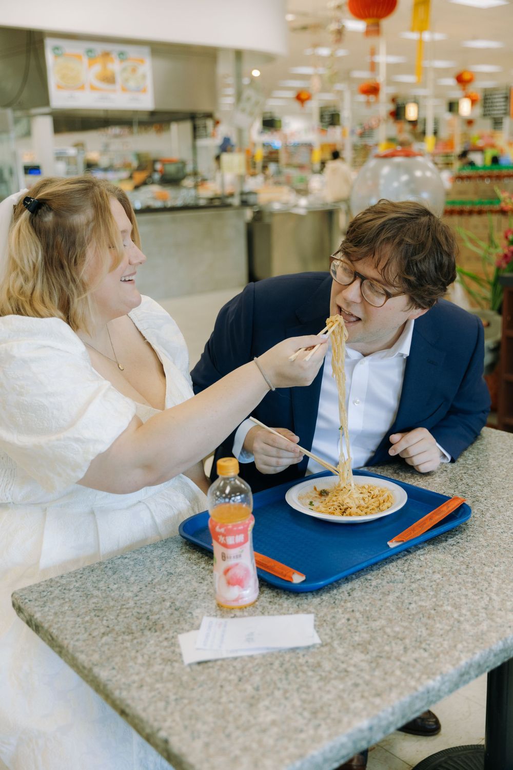 A couple shares a playful moment while eating spaghetti at a casual restaurant with festive decorations in the background.