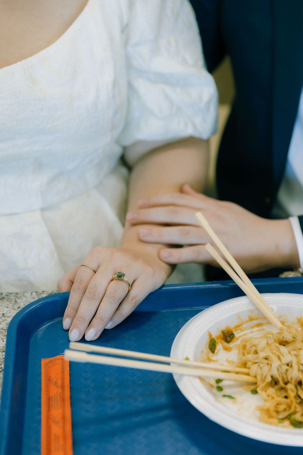 Person wearing white blouse and emerald ring uses chopsticks to eat noodles from a plate on a blue cafeteria tray.
