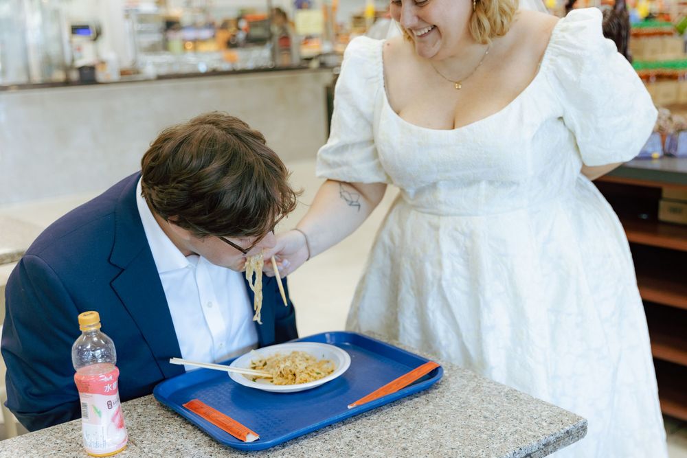 A couple in wedding attire share a laugh at a cafeteria while one eats from a tray.