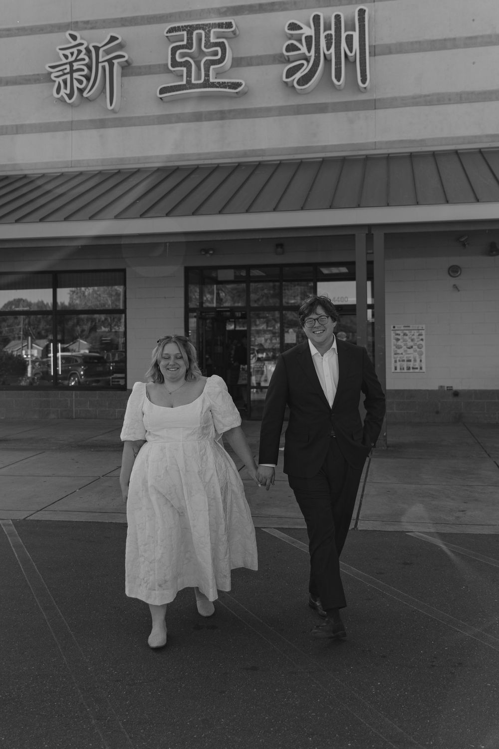 A couple walks together outside an Asian restaurant storefront in a black and white photograph.