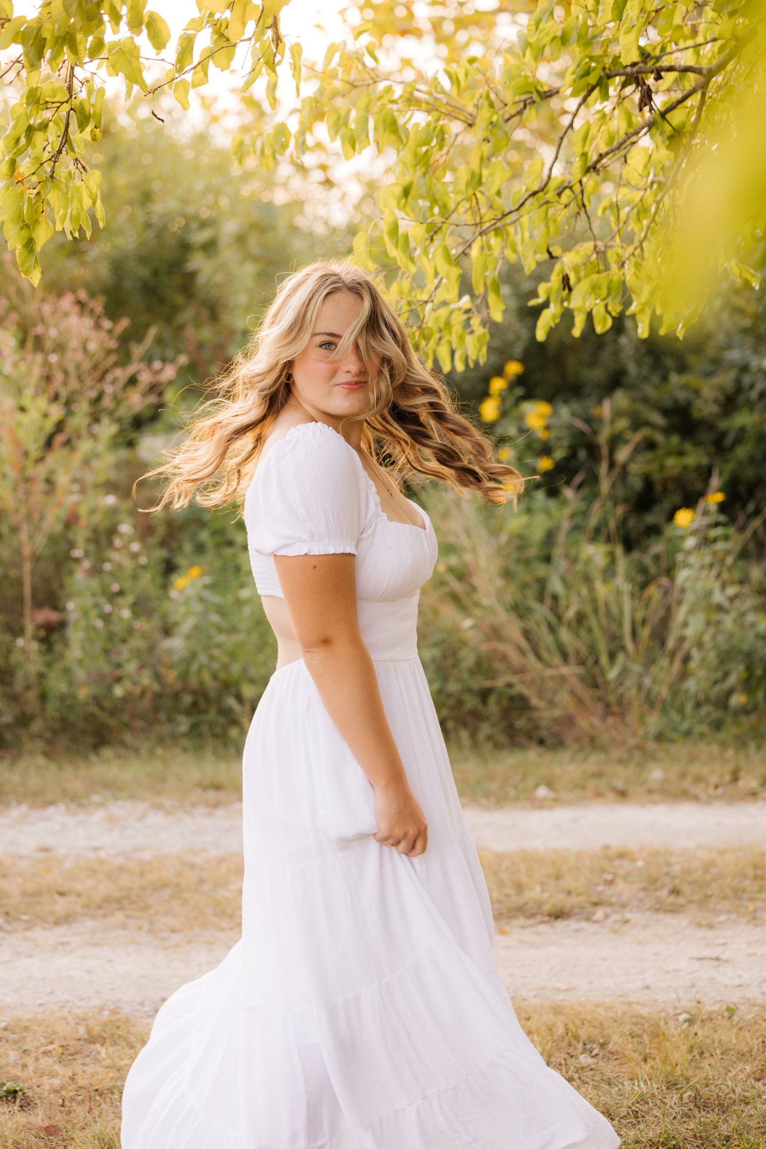 Young woman in flowing white dress twirls amid golden autumn foliage with sunlight glowing through yellow leaves.