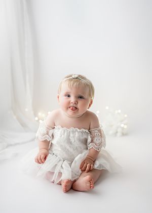 Baby in white lace dress sits on white backdrop with twinkle lights behind.