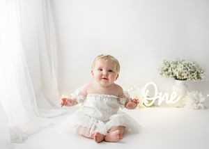 Baby in white dress sits on floor with illuminated 'One' sign and white flowers in background for first birthday photoshoot.