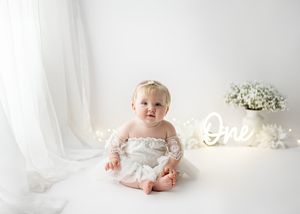 Baby in white lace dress sits on white backdrop with illuminated 'One' sign and baby's breath flowers for first birthday photo.