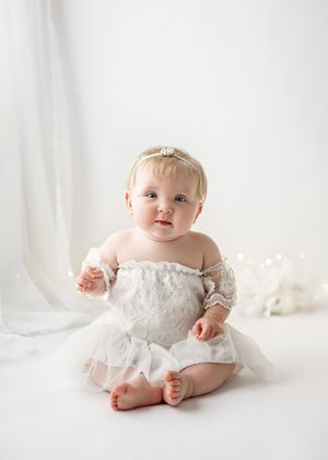 Baby wearing white lace dress sits against light backdrop during studio portrait session.