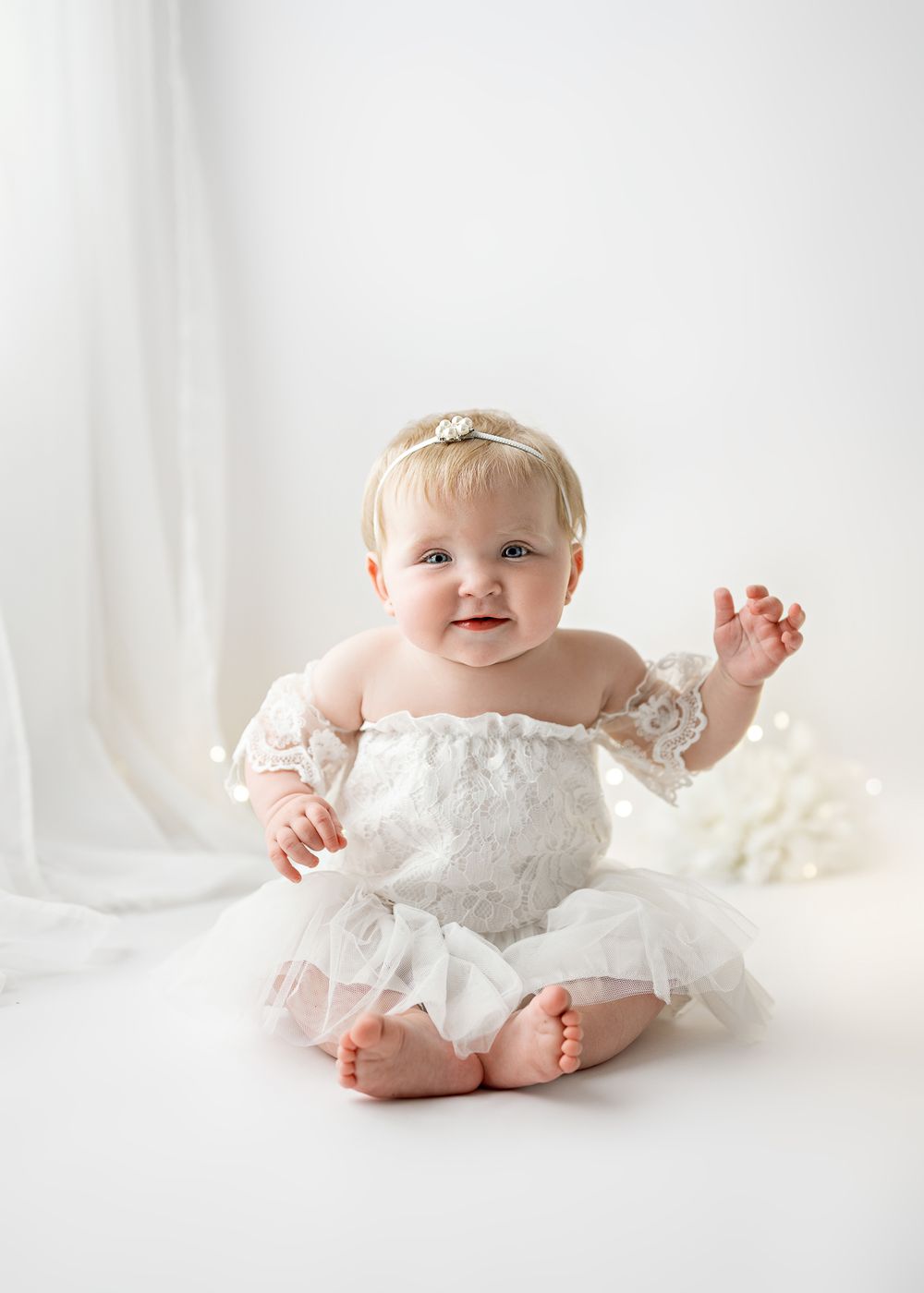 A baby wearing a delicate white lace dress sits on soft white fabric in a bright minimalist setting.