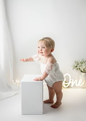 Baby in white lace romper stands next to white cube and illuminated 'One' sign in minimalist studio setting.