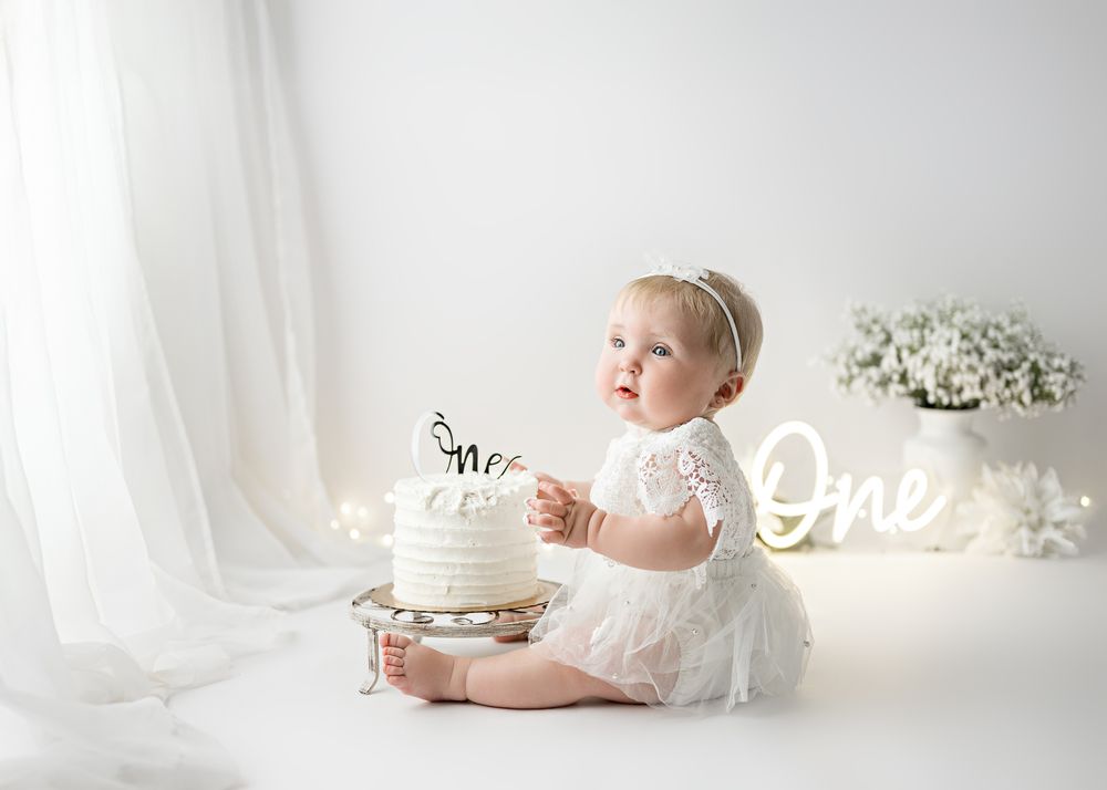 A baby in white lace dress sits by a decorated cake against a light background with neon 'One' sign and flowers.