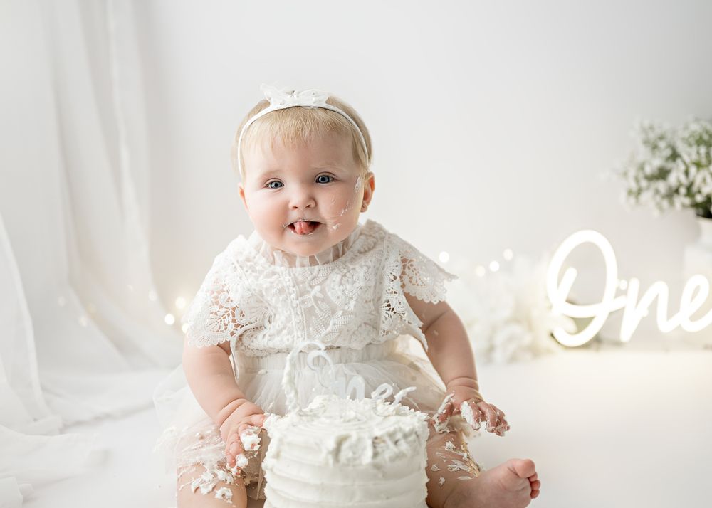 Baby in white lace dress sits next to glowing neon 'One' sign and smashes white birthday cake against light backdrop.