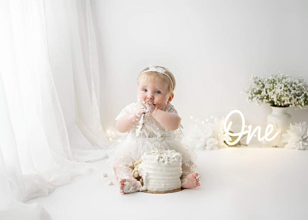 Sweet first birthday cake smash photo session with white cake and glowing 'One' sign in background.