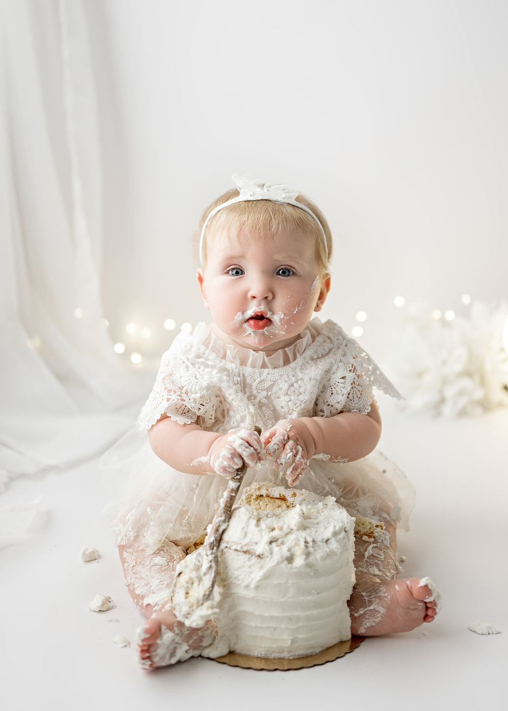 A baby in a white lace dress sits on a white backdrop with twinkle lights while playing with a white cake.