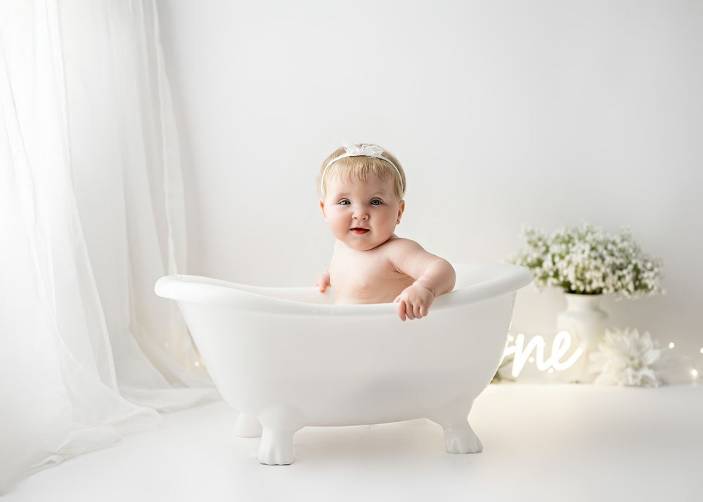 A baby sits in a white vintage bathtub with a neon 'One' sign and florals in a bright studio setting.