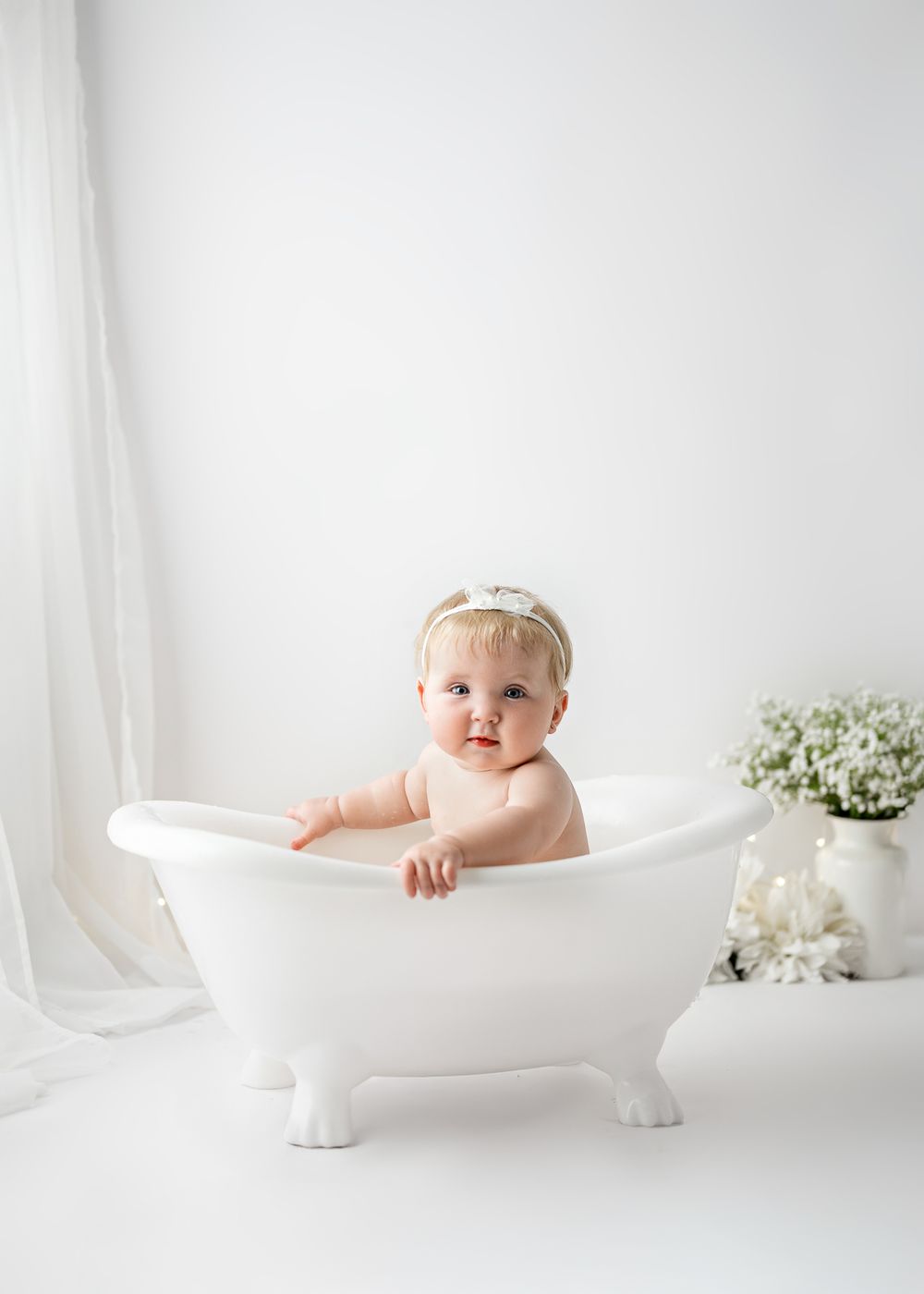 Baby sitting in vintage white clawfoot bathtub with soft white curtain backdrop and floral decor.