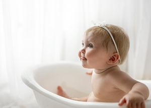 A baby wearing a headband sits in a white bathtub looking upward against a light background.