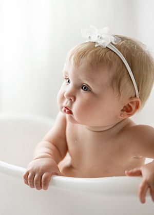 Baby wearing a white bow headband peers over the edge of a bathtub during bathtime.