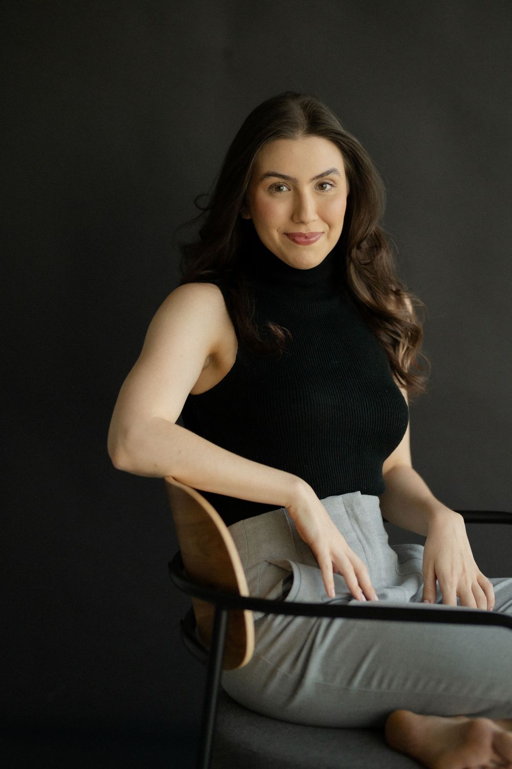 A young woman with long dark hair sits relaxed in a chair, wearing a black turtleneck top and gray pants.