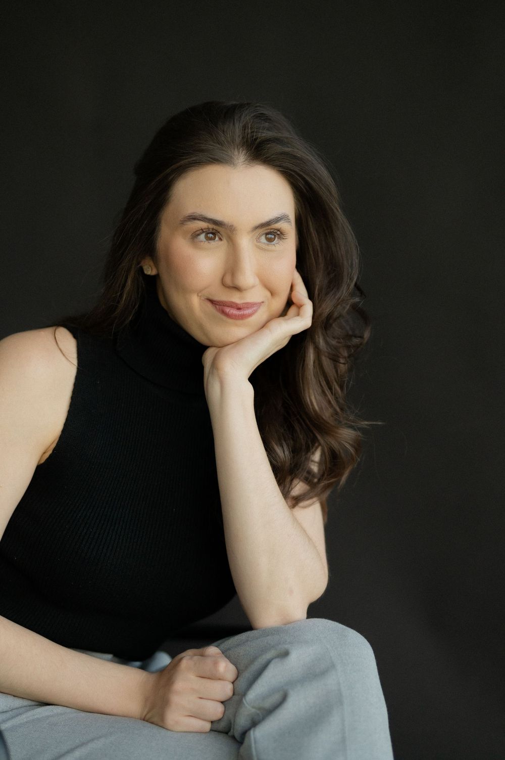 Young woman in black turtleneck and gray pants poses confidently with hand on chin against dark background.