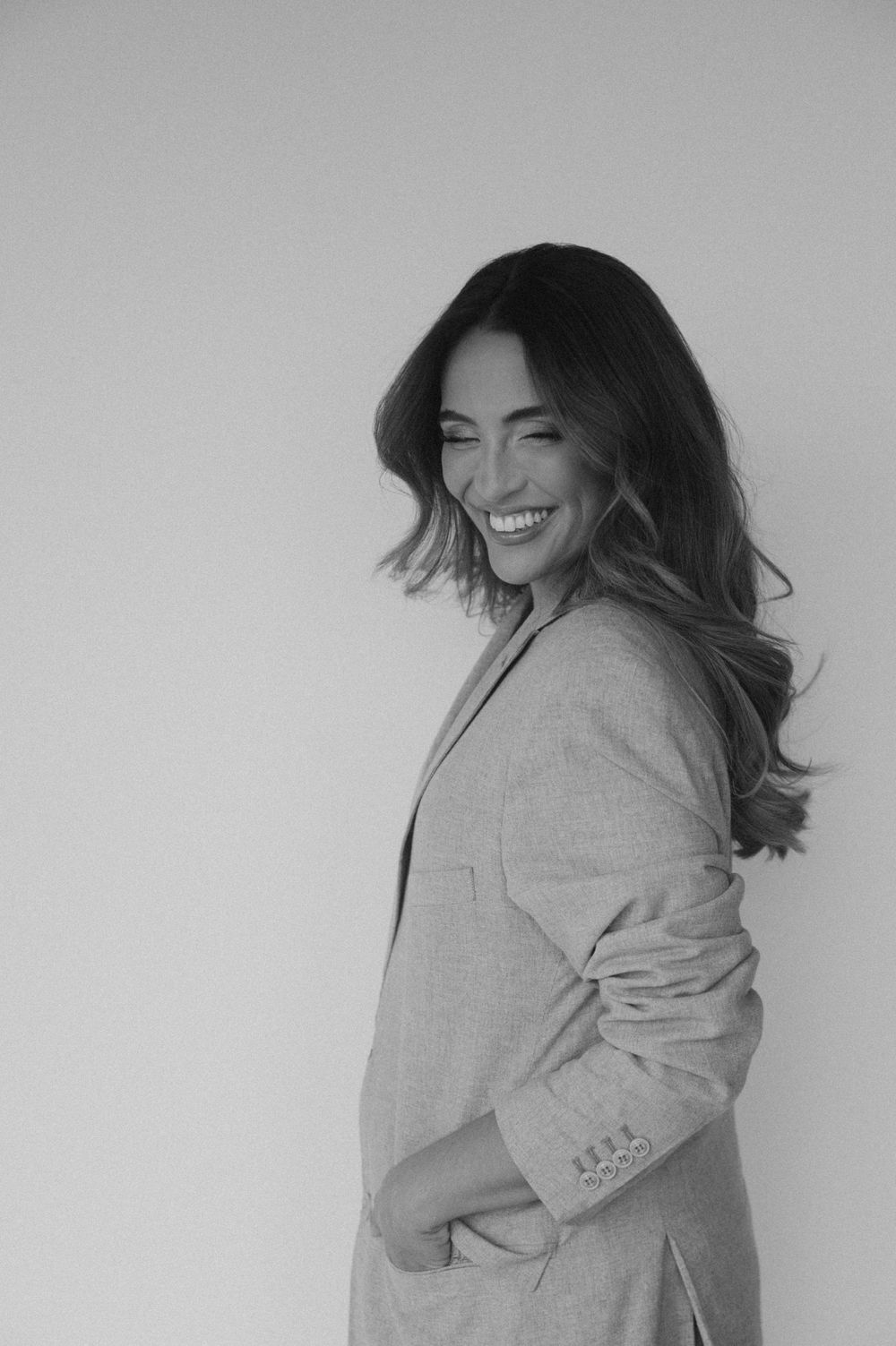 Black and white portrait of a smiling woman with long hair wearing a blazer, posing confidently against a light background.