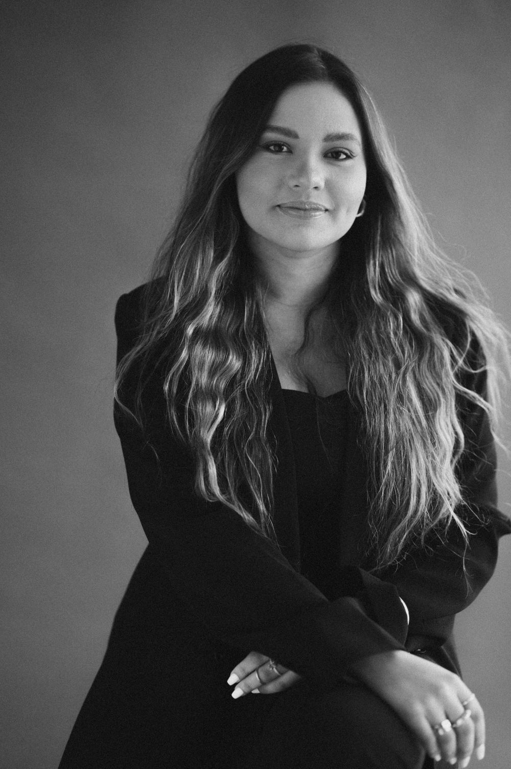 Woman in black blazer with long wavy hair poses confidently in a professional black and white portrait photo.