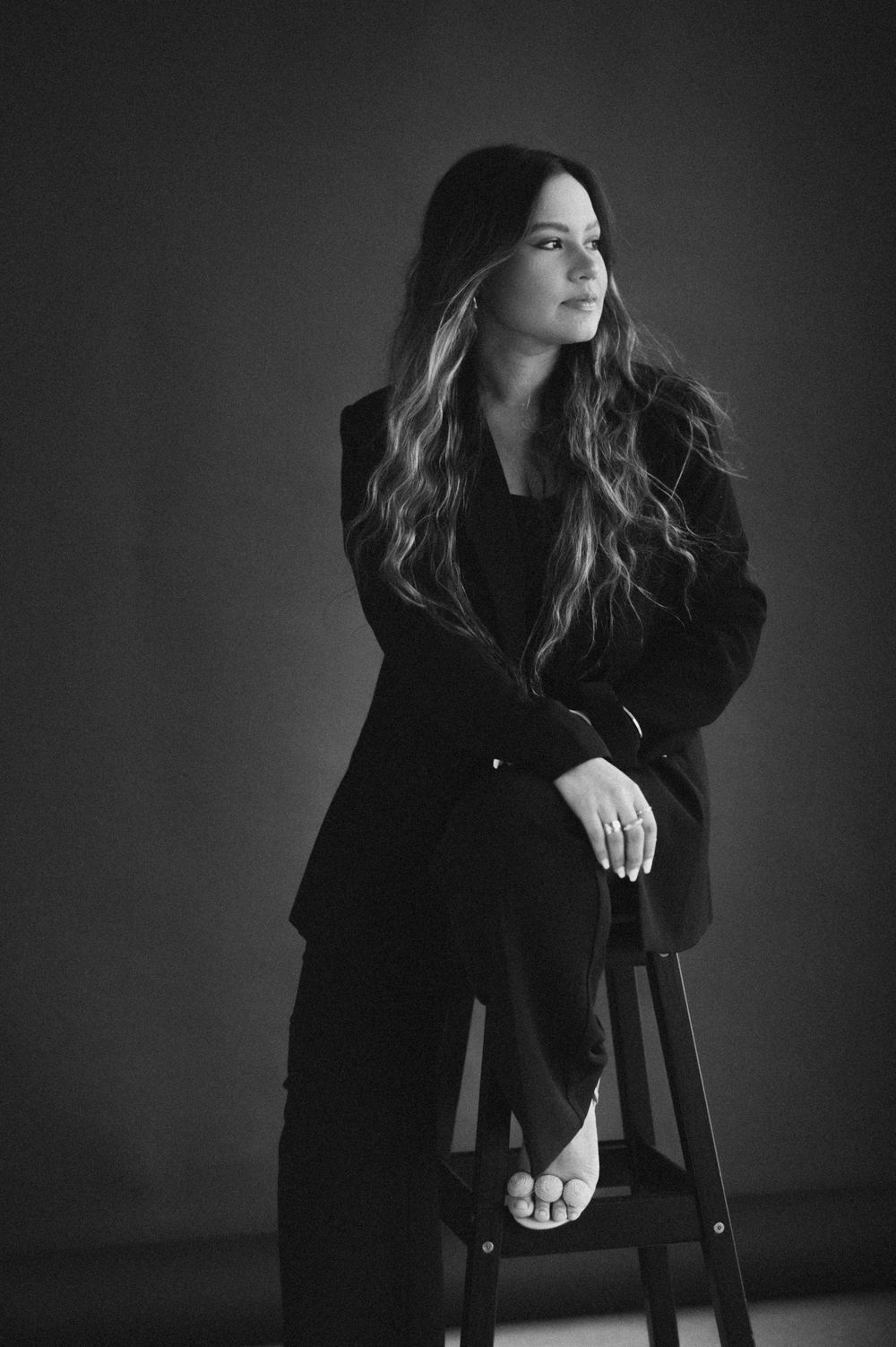 Woman in black suit sitting on stool, black and white studio portrait with dramatic lighting.