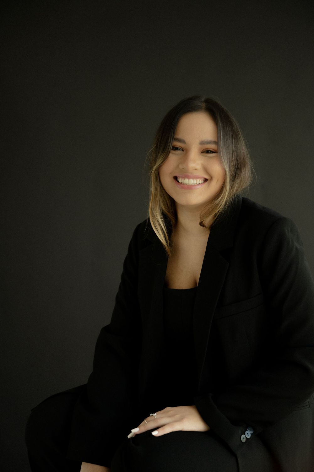 Smiling young woman in black blazer poses confidently against dark background in professional portrait photo.