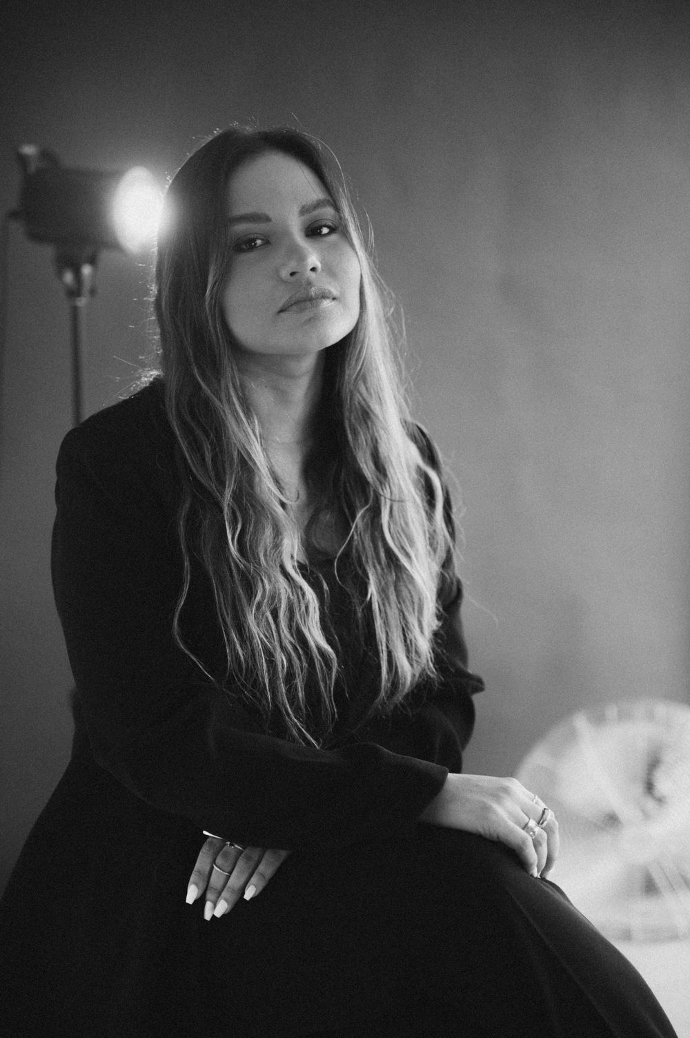 Black and white portrait of a woman with long wavy hair in a black outfit, seated with studio light behind her.