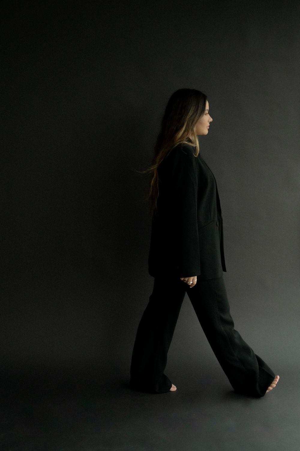 Woman in black oversized blazer and wide-leg pants walking against dark studio background.