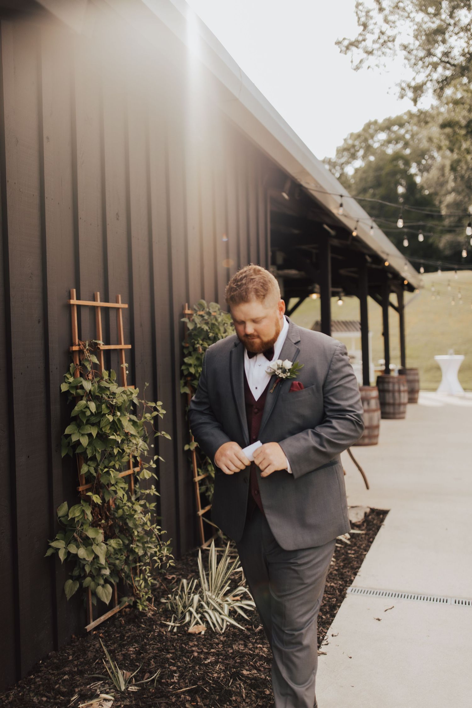 A person in a grey suit and bowtie adjusts their outfit while standing outside a black barn-style building on a sunny day.