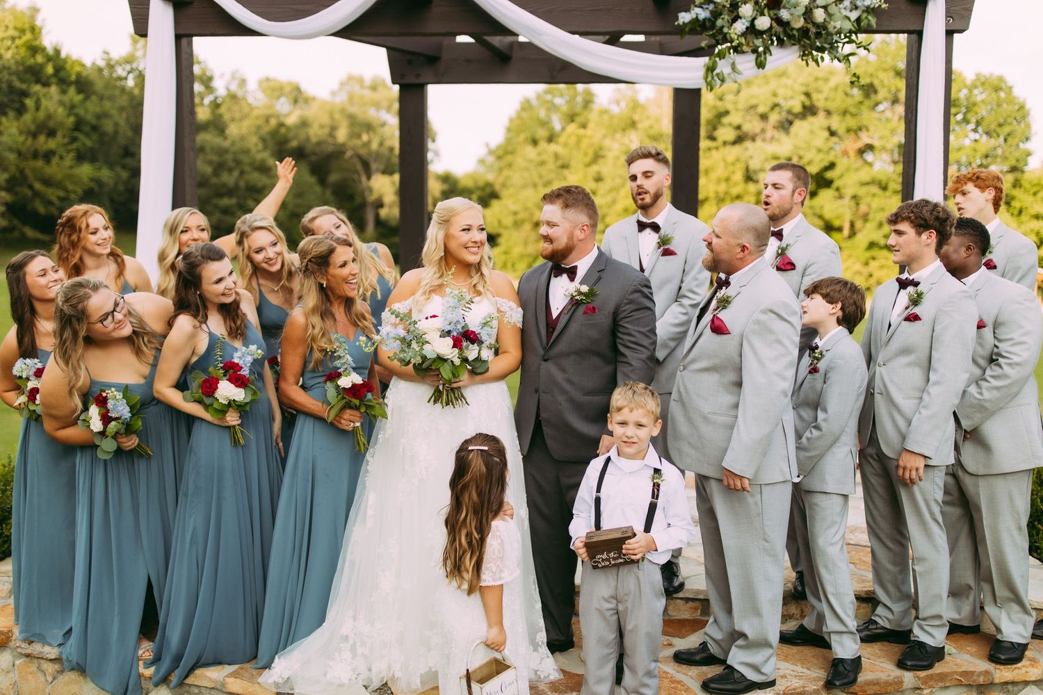 Wedding party in teal bridesmaid dresses and gray suits poses together outdoors under a gazebo during summer ceremony.