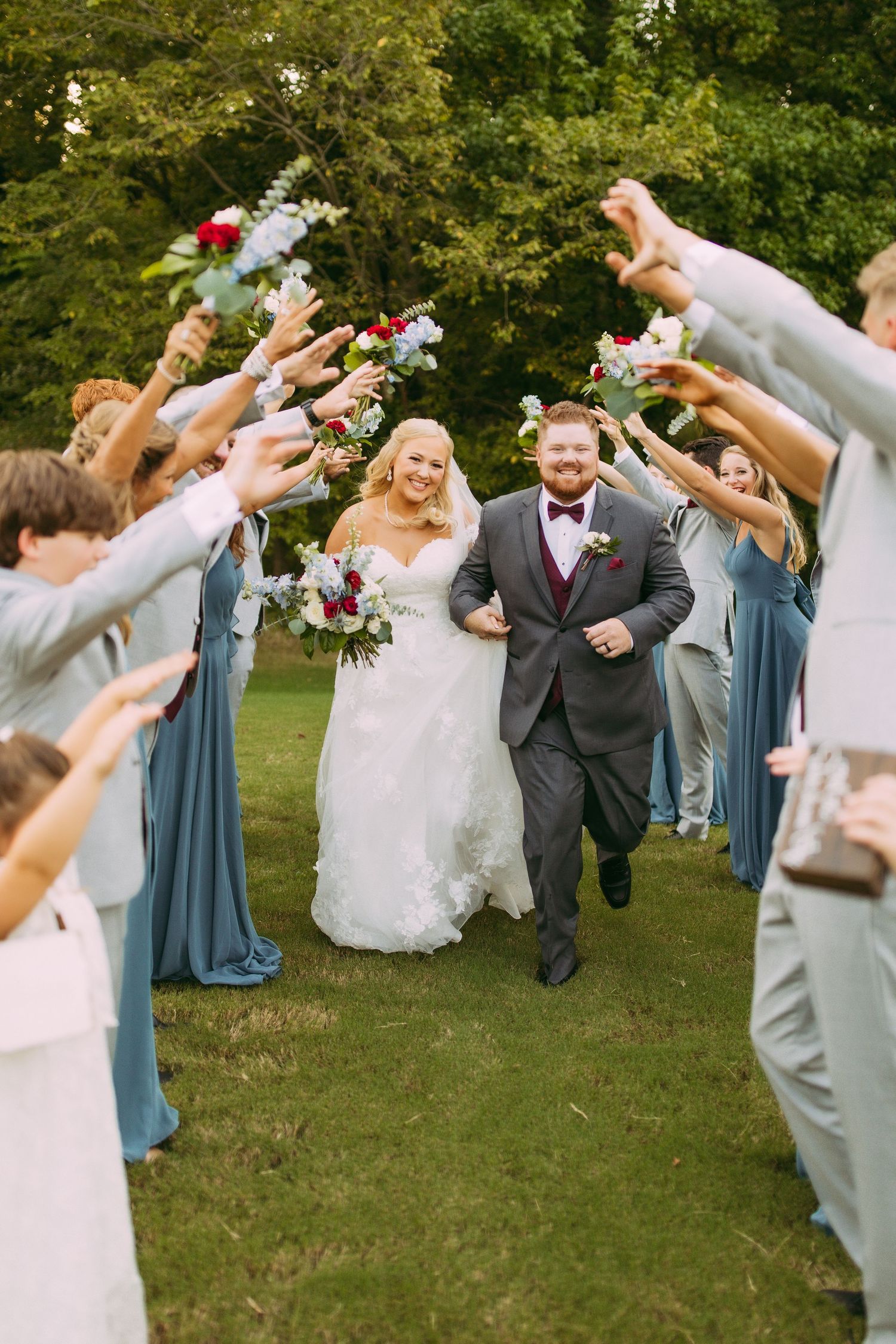 Wedding party members form an aisle on grassy lawn, raising their arms in celebration as newlyweds walk through.