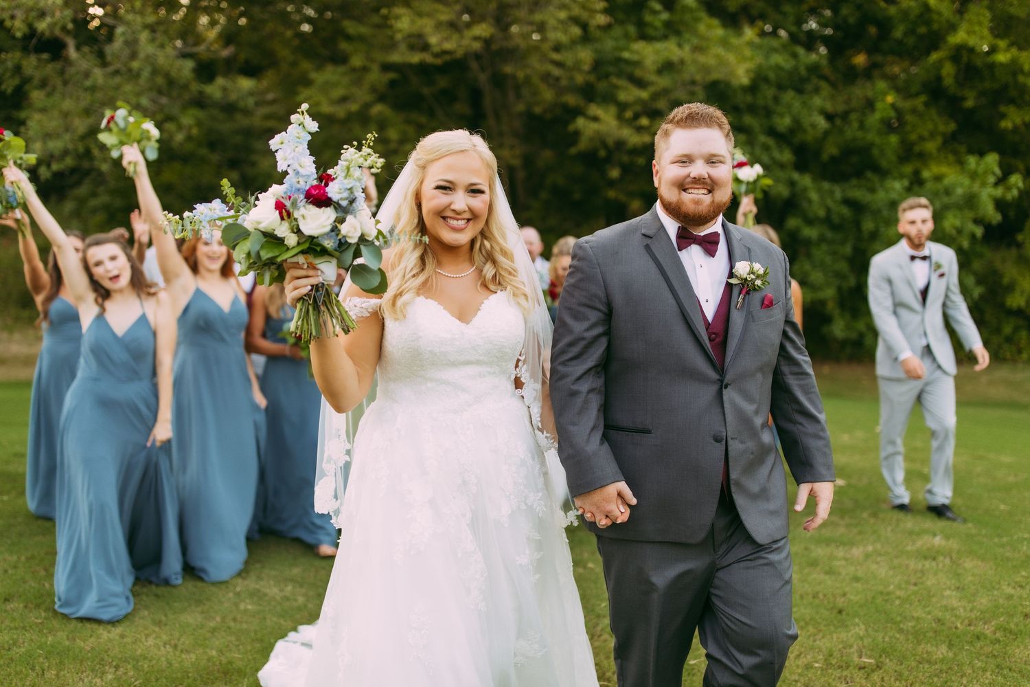 Wedding party celebrates outdoors with bridesmaids in blue dresses cheering behind the newlyweds on grassy lawn.