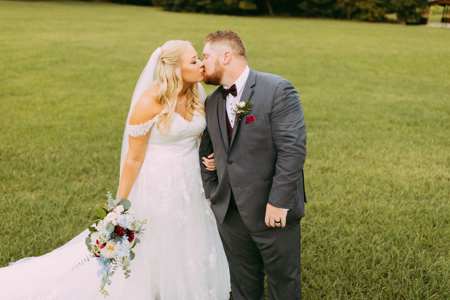 A couple shares a tender moment on their wedding day in a green field while holding a rustic floral bouquet.