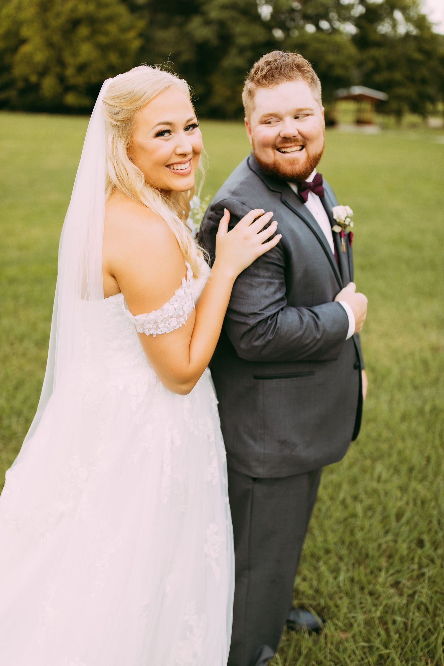 A bride in a white lace gown with veil poses with groom in charcoal suit against a lush green outdoor setting.