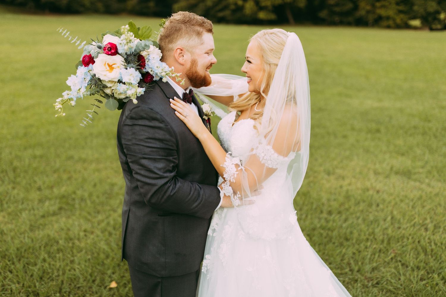 A loving couple embraces outdoors on their wedding day, with the bride holding a colorful bouquet in a grassy field.