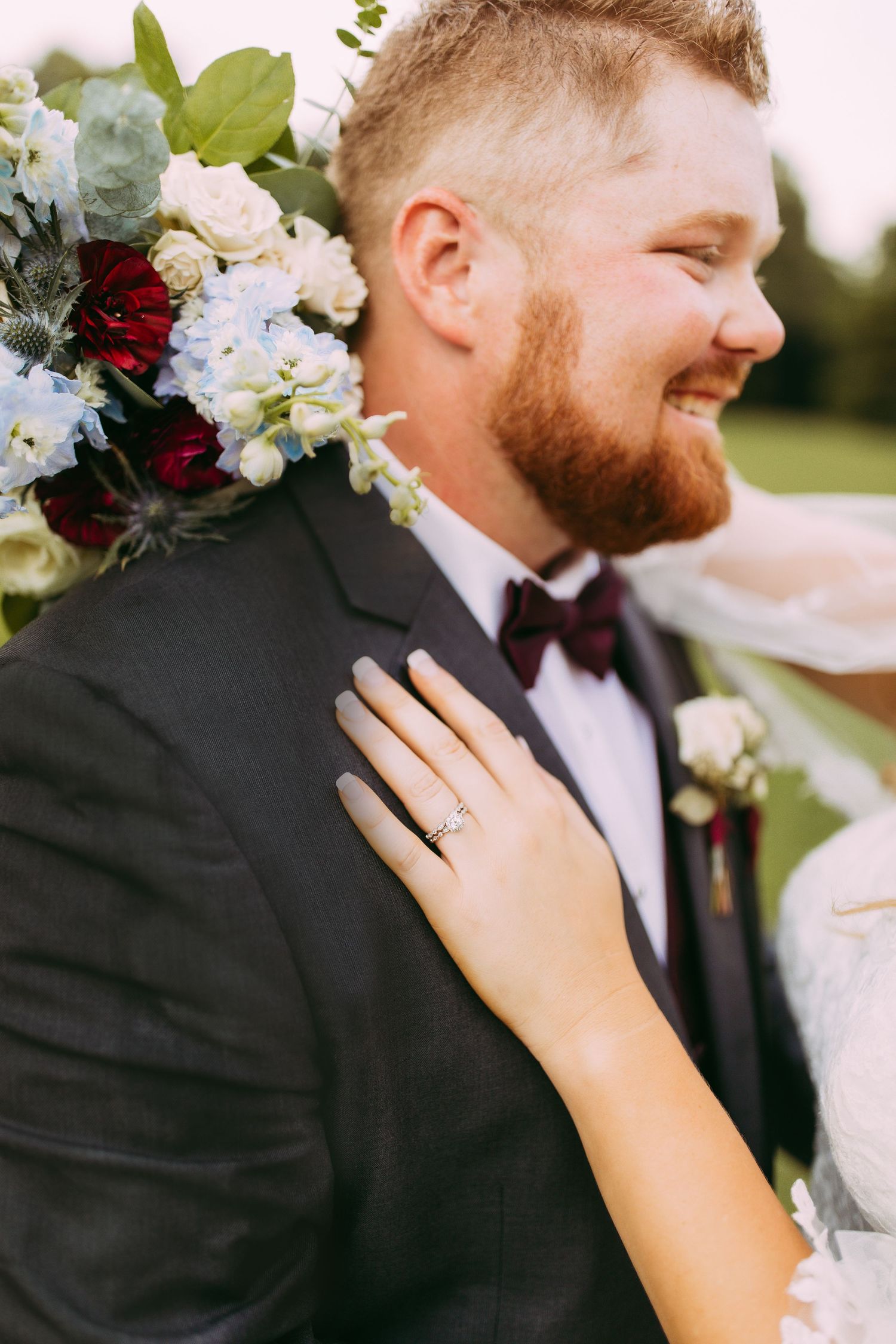 Candid outdoor wedding photograph showing a groom in classic tuxedo and burgundy bow tie with a lush floral bouquet.