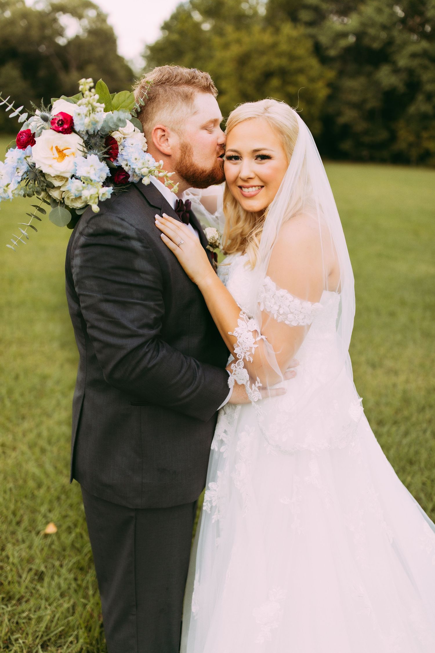 Newlyweds share tender moment outdoors with bride in white gown holding floral bouquet and groom in black suit.