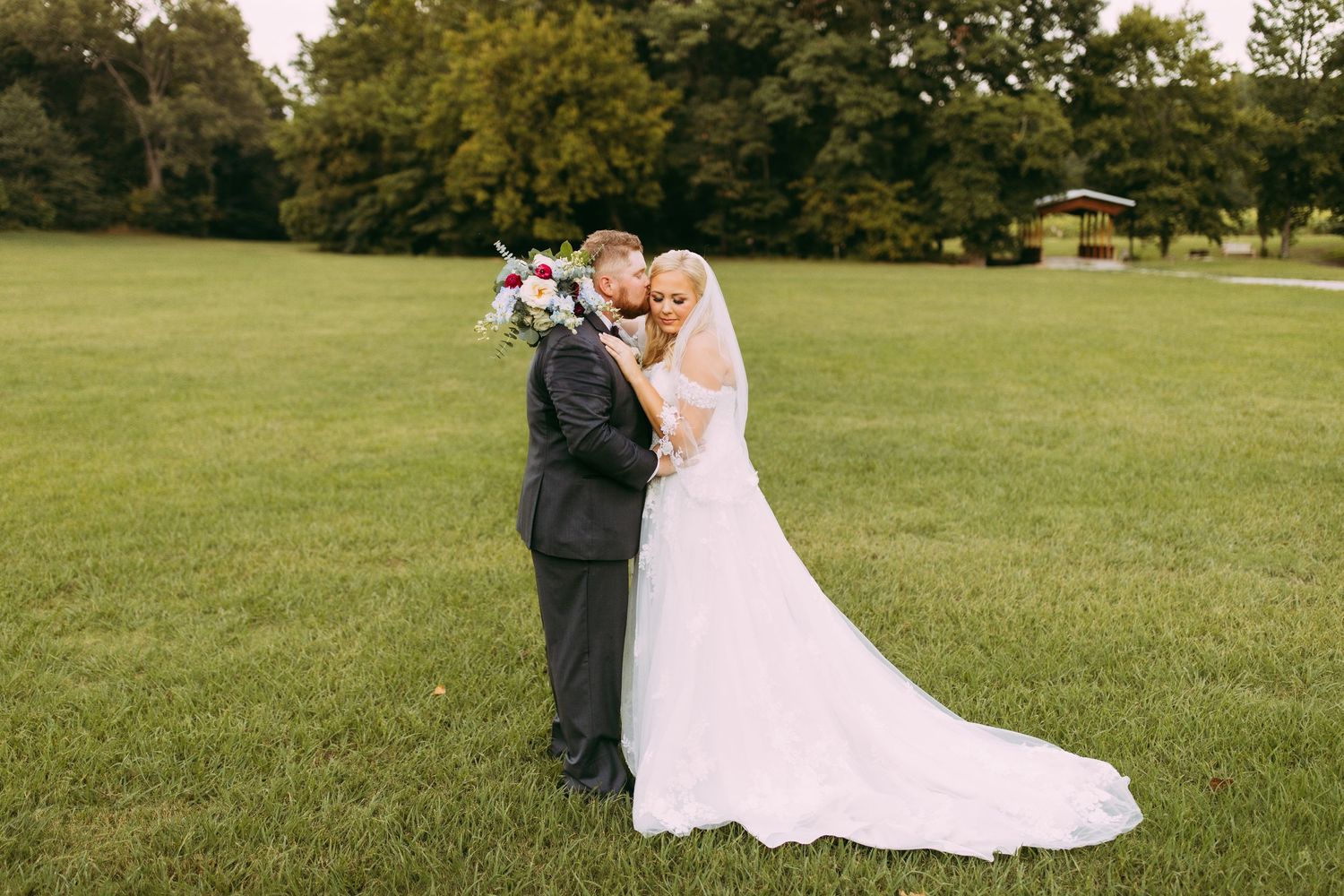 A romantic wedding moment captured on a lush green lawn with trees in the background during an outdoor ceremony.