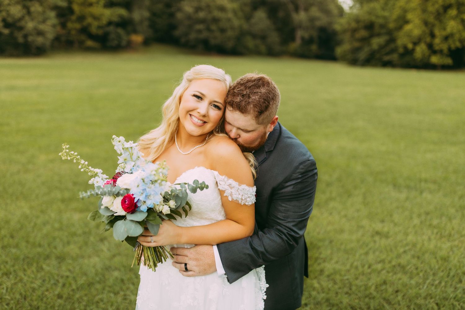 A newlywed couple embraces in a sunlit green field as the bride holds a bouquet of purple and white flowers.