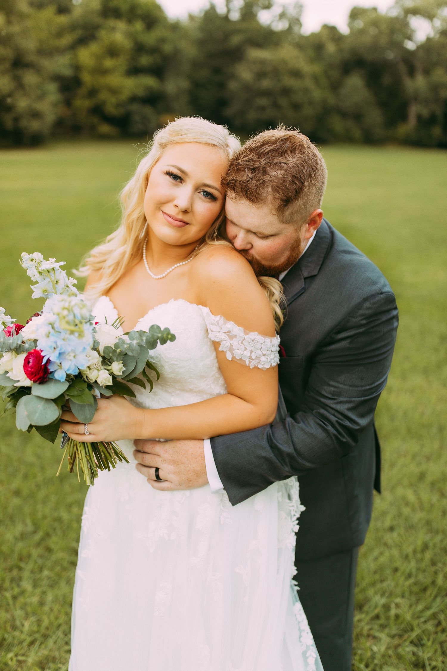 A romantic outdoor wedding photo showing a couple embracing in a green field with a colorful bridal bouquet.