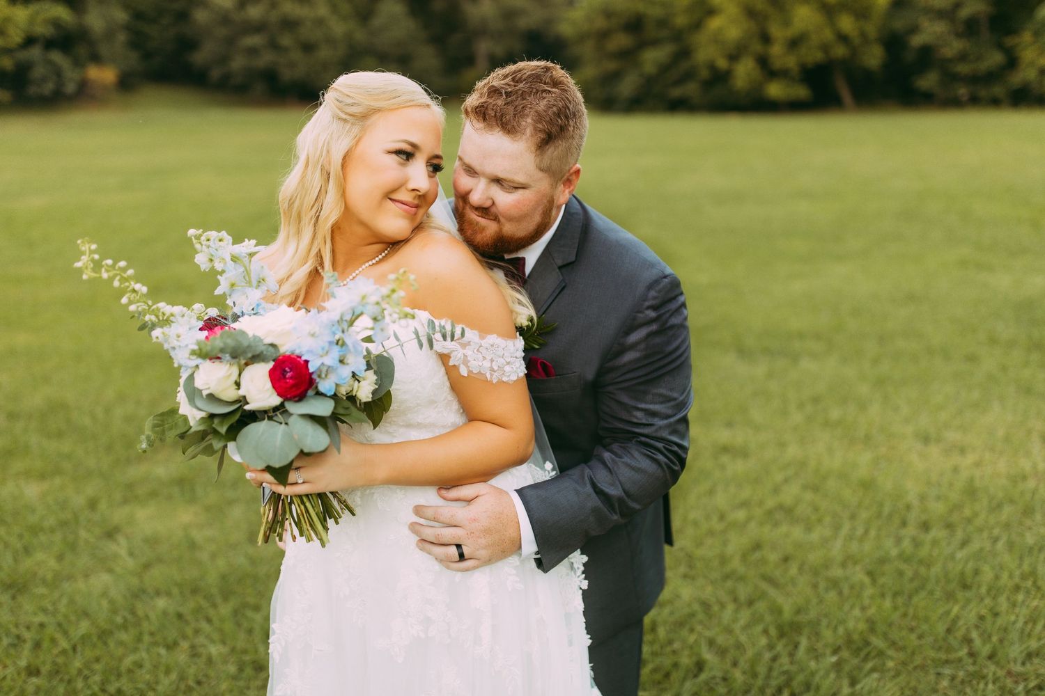 A newlywed couple embraces on a grassy lawn while holding a beautiful bouquet of pink and purple flowers.