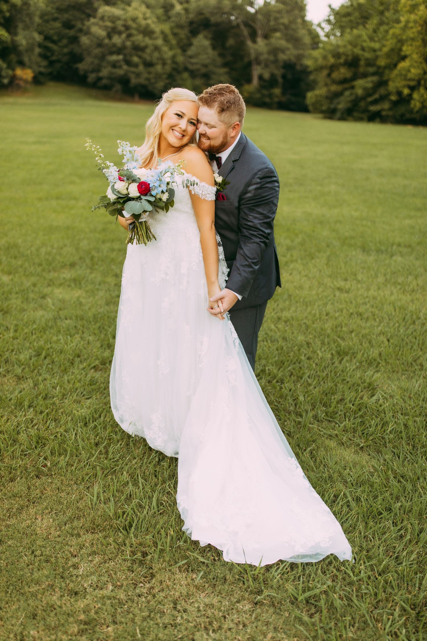 Newlyweds share an intimate moment on a green lawn while the bride holds a purple and white floral bouquet.