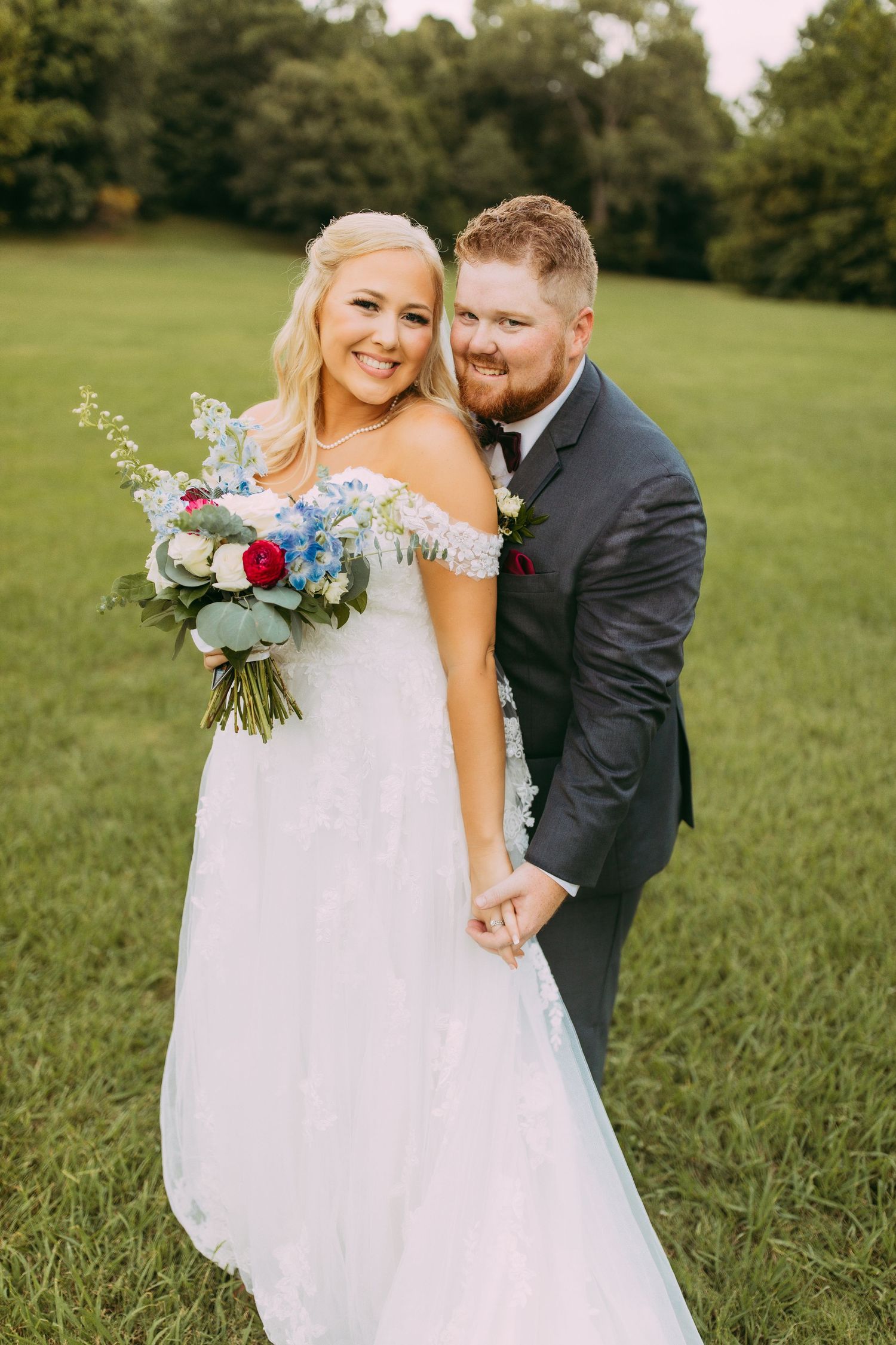 A newlywed couple poses in a green field with the bride holding a beautiful bouquet of blue and red flowers.