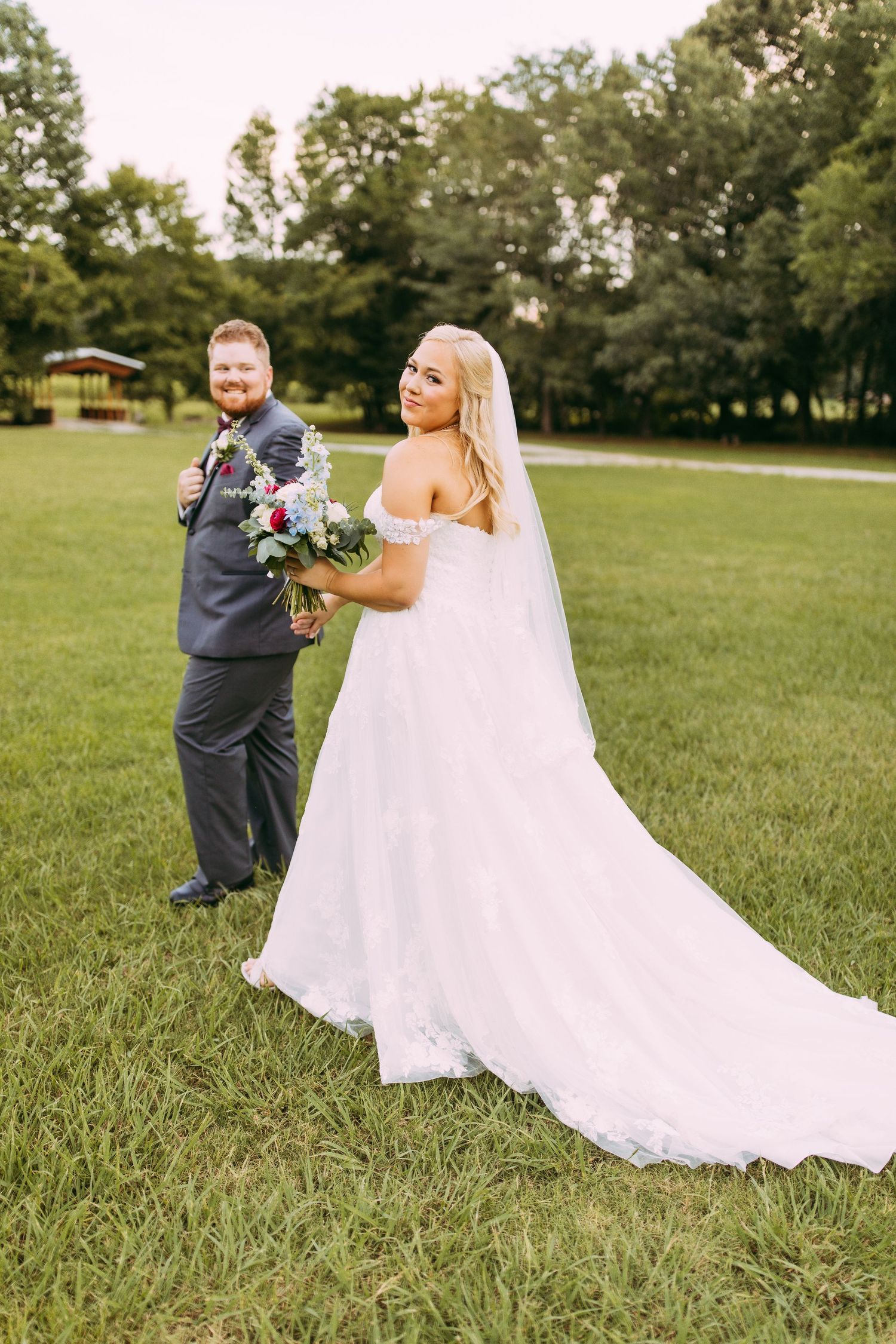 A couple in wedding attire stands on a grassy lawn with trees and a gazebo in the background.