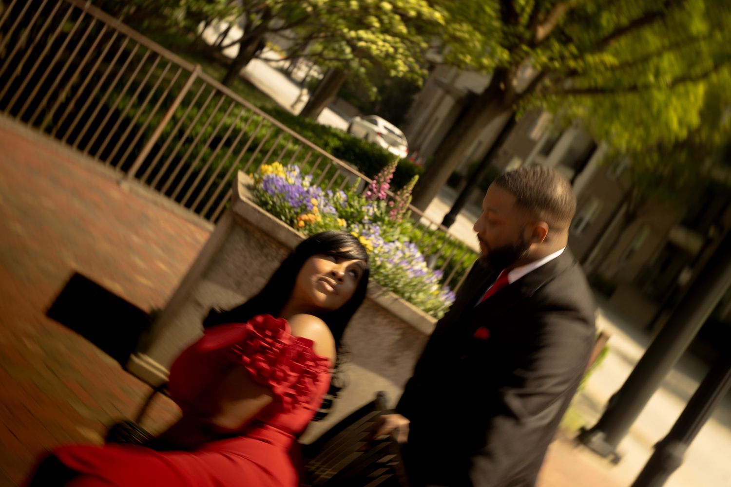 A couple in formal attire stands outside on a brick walkway with someone in a red dress and another in a dark suit.