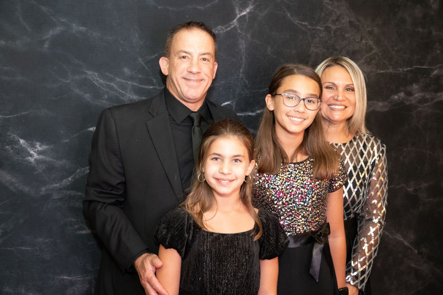 A family of four poses for a formal portrait against a dark marble backdrop.
